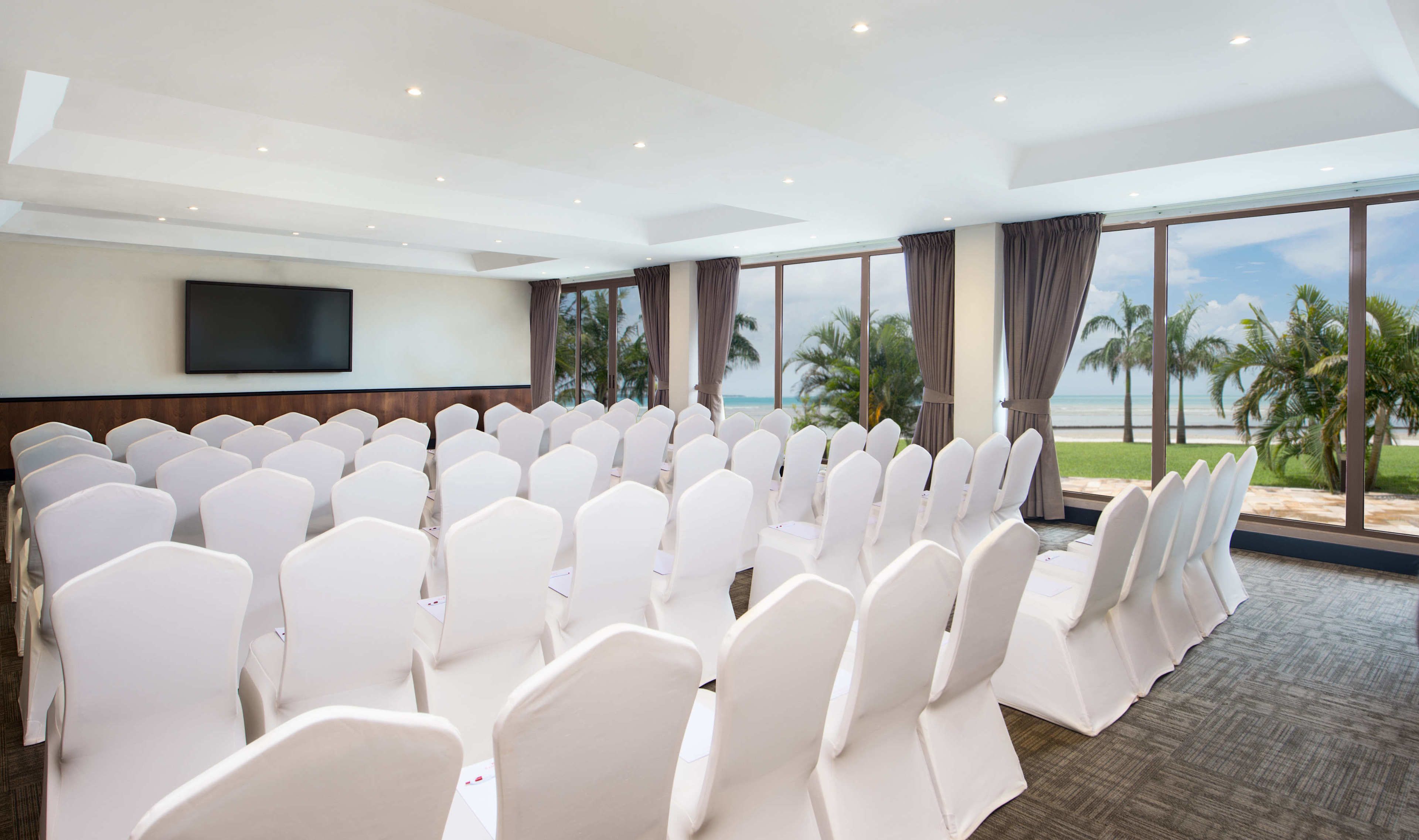 A conference room with rows of white chairs, a flat-screen TV on the wall, and large windows offering a view of the beach.