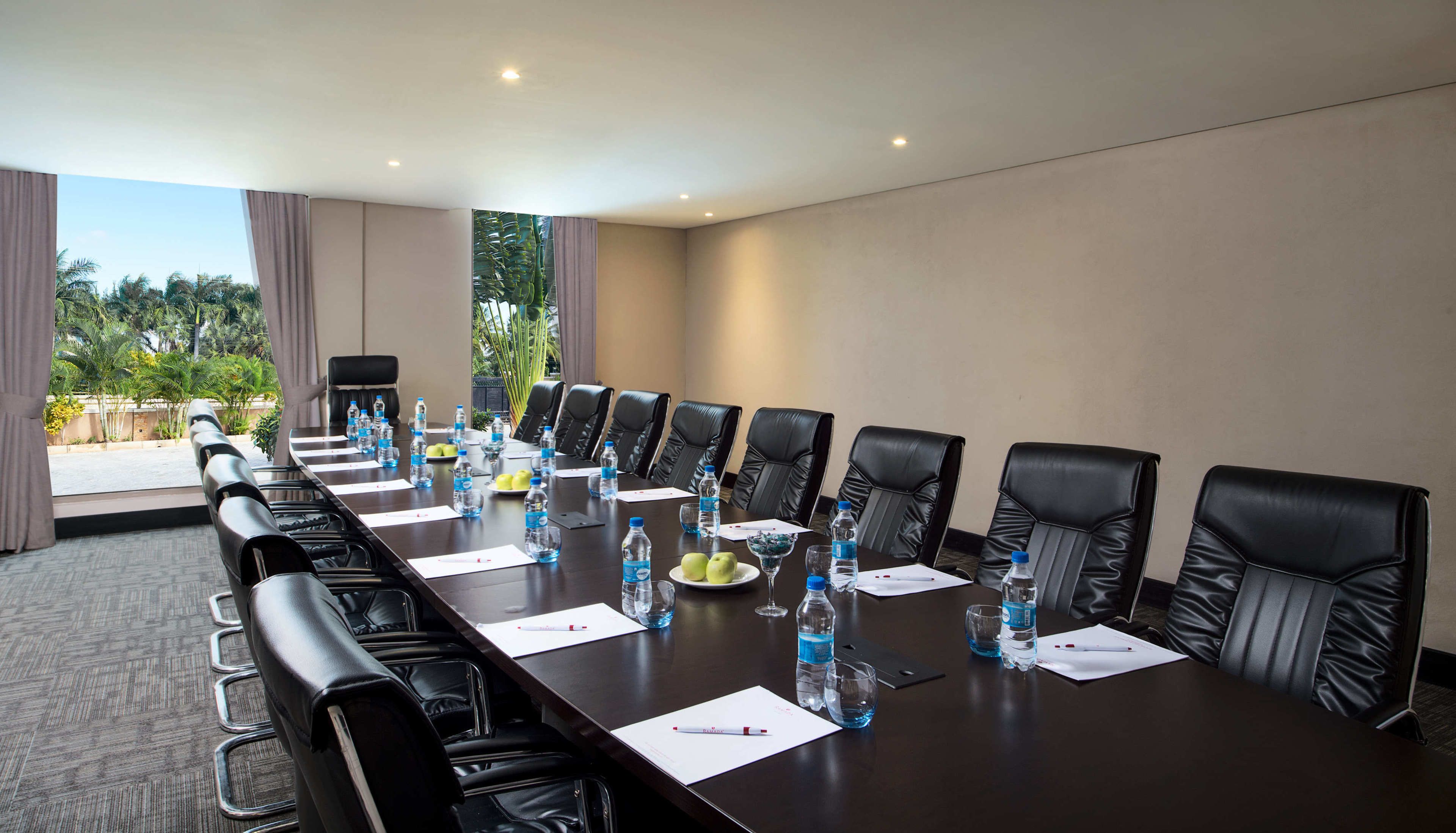 A conference room with a long table, black leather chairs, water bottles, and a view of palm trees.