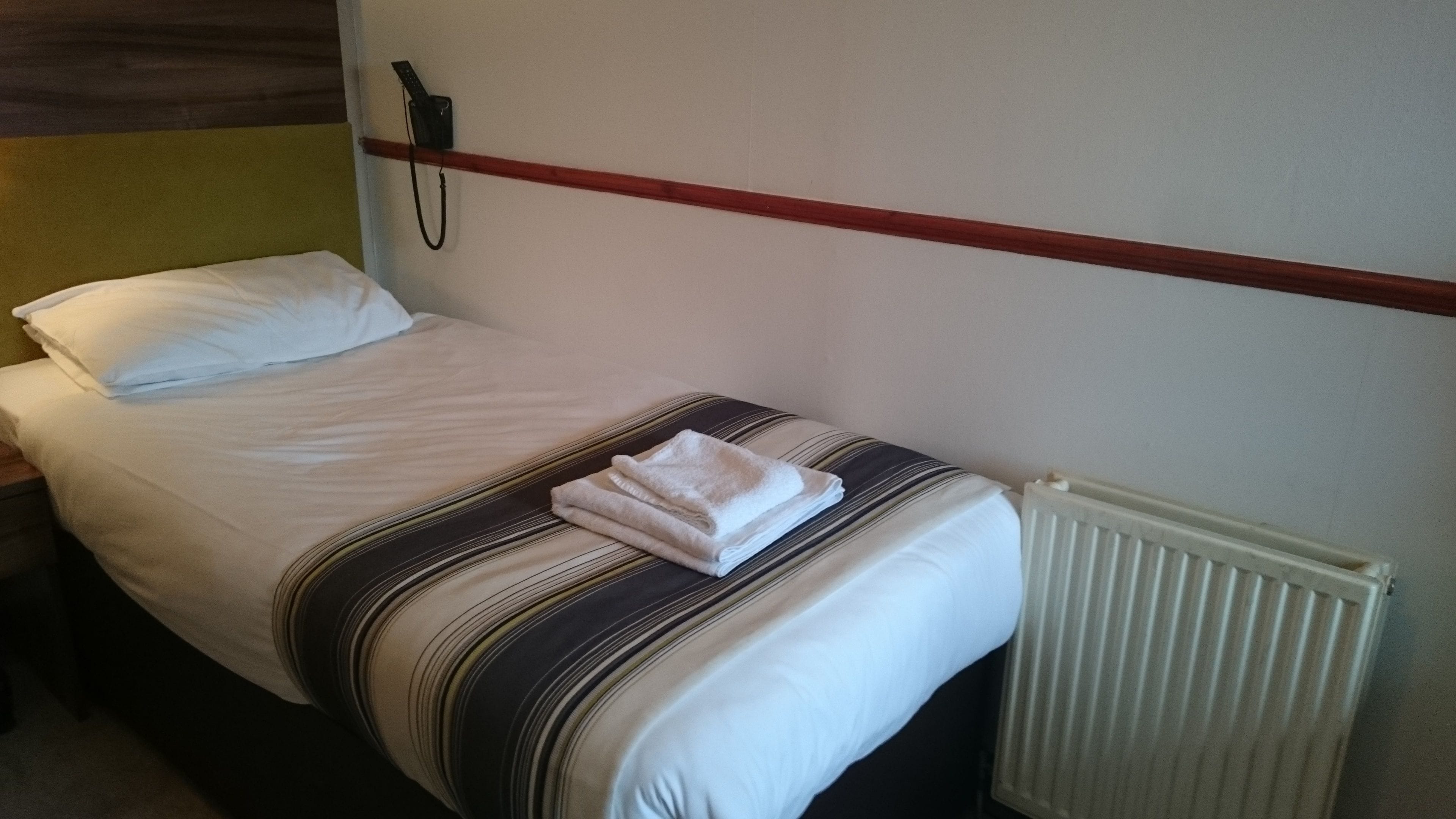 A single bed with a striped blanket, a white pillow, and a folded towel. There is a telephone on the wall to the left of the bed.