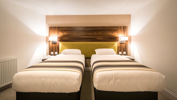 A hotel room with two single beds, a wooden headboard, bedside lamps, and a radiator. - Livingston Lodge Hotel (Livingston)