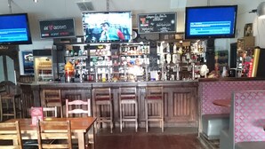 A bar area with wooden tables and chairs, a counter with various bottles and glasses, and two televisions displaying content. - Livingston Lodge Hotel (Livingston)