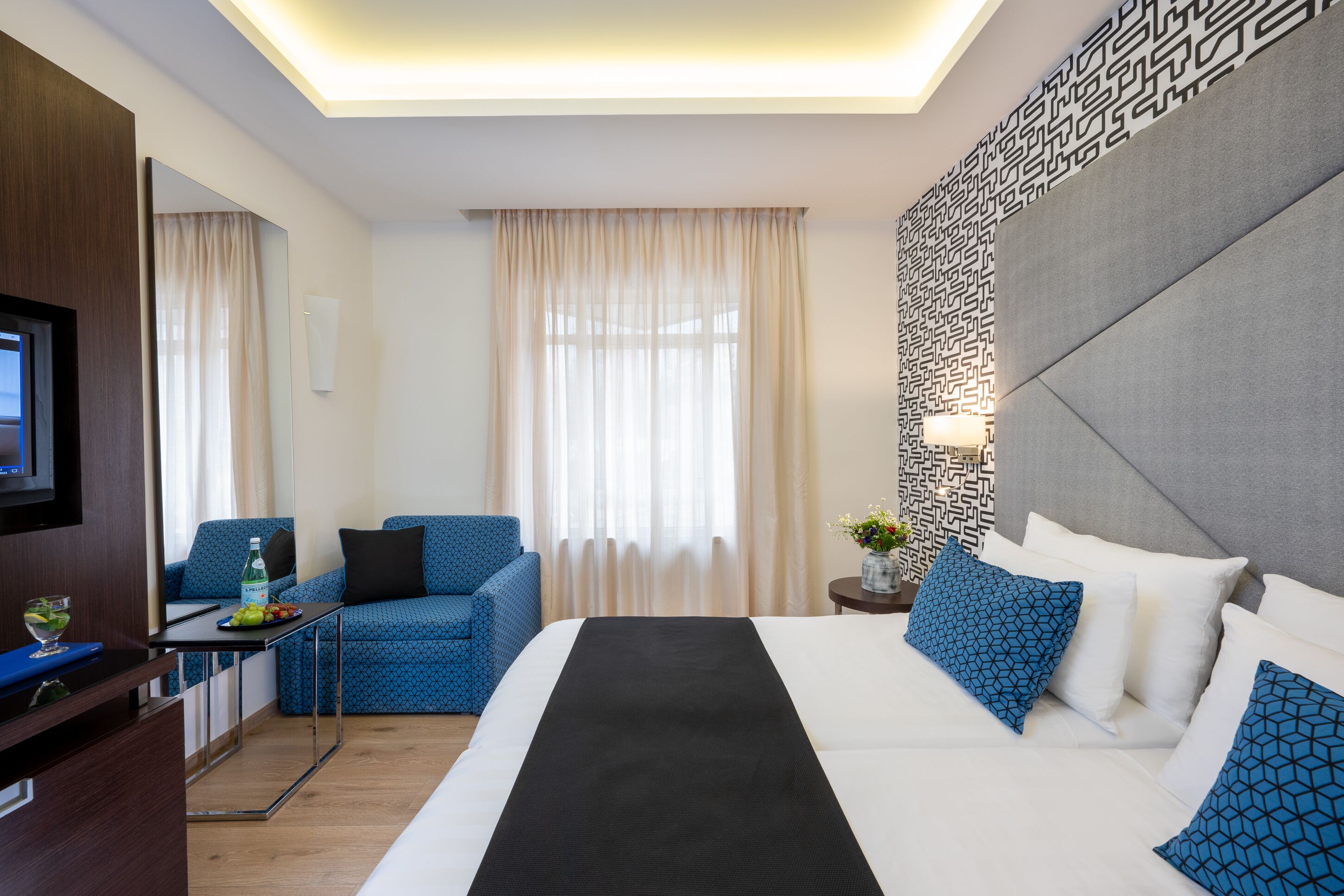 A hotel room with a bed, a sofa, a TV, and a small table with a bottle and fruit.