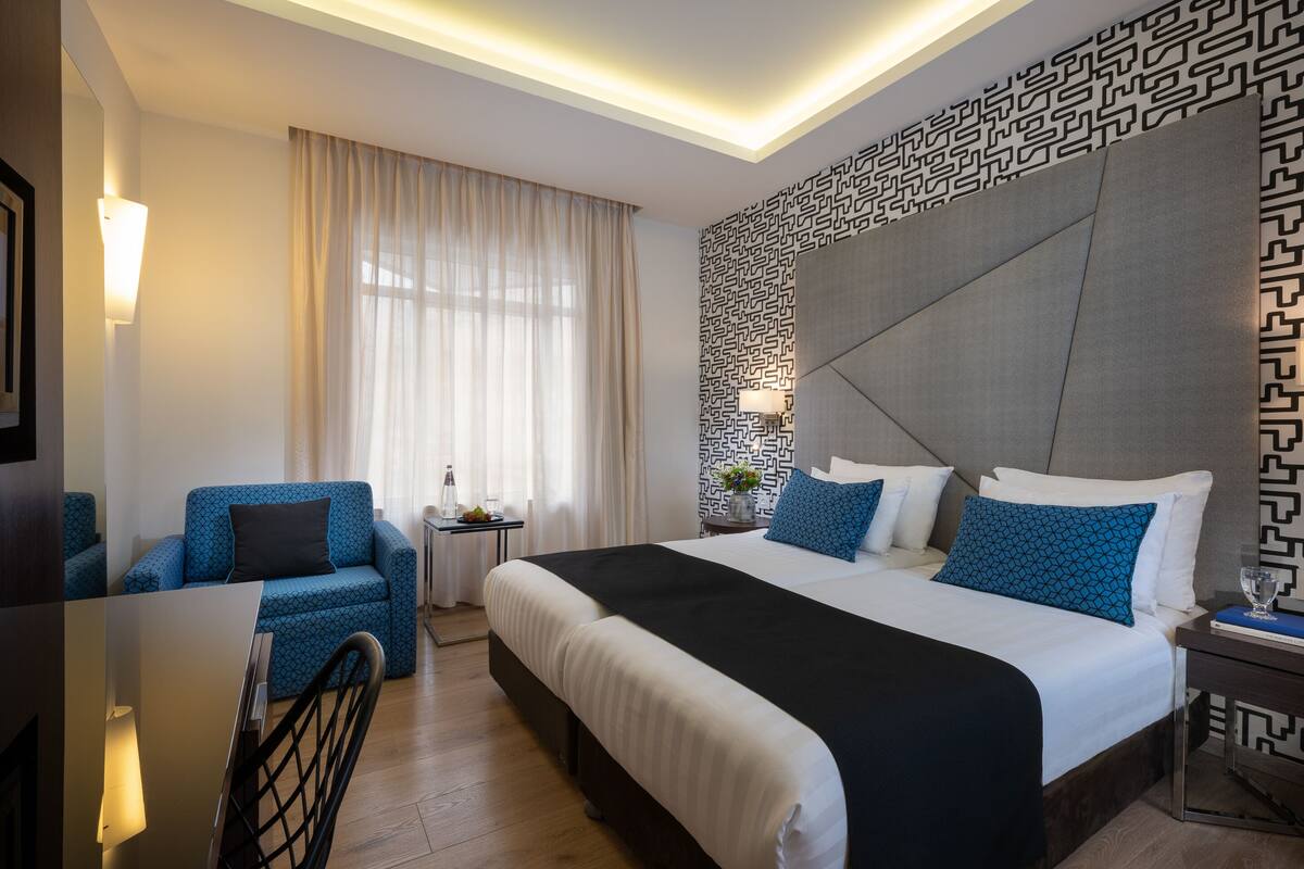 Habitación de hotel con una cama grande, un sillón azul, una mesita de noche con lámpara y un papel tapiz estampado.