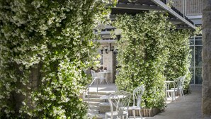 Een overdekt terras met witte metalen stoelen en tafels, omgeven door weelderige begroeiing en stenen vloeren.