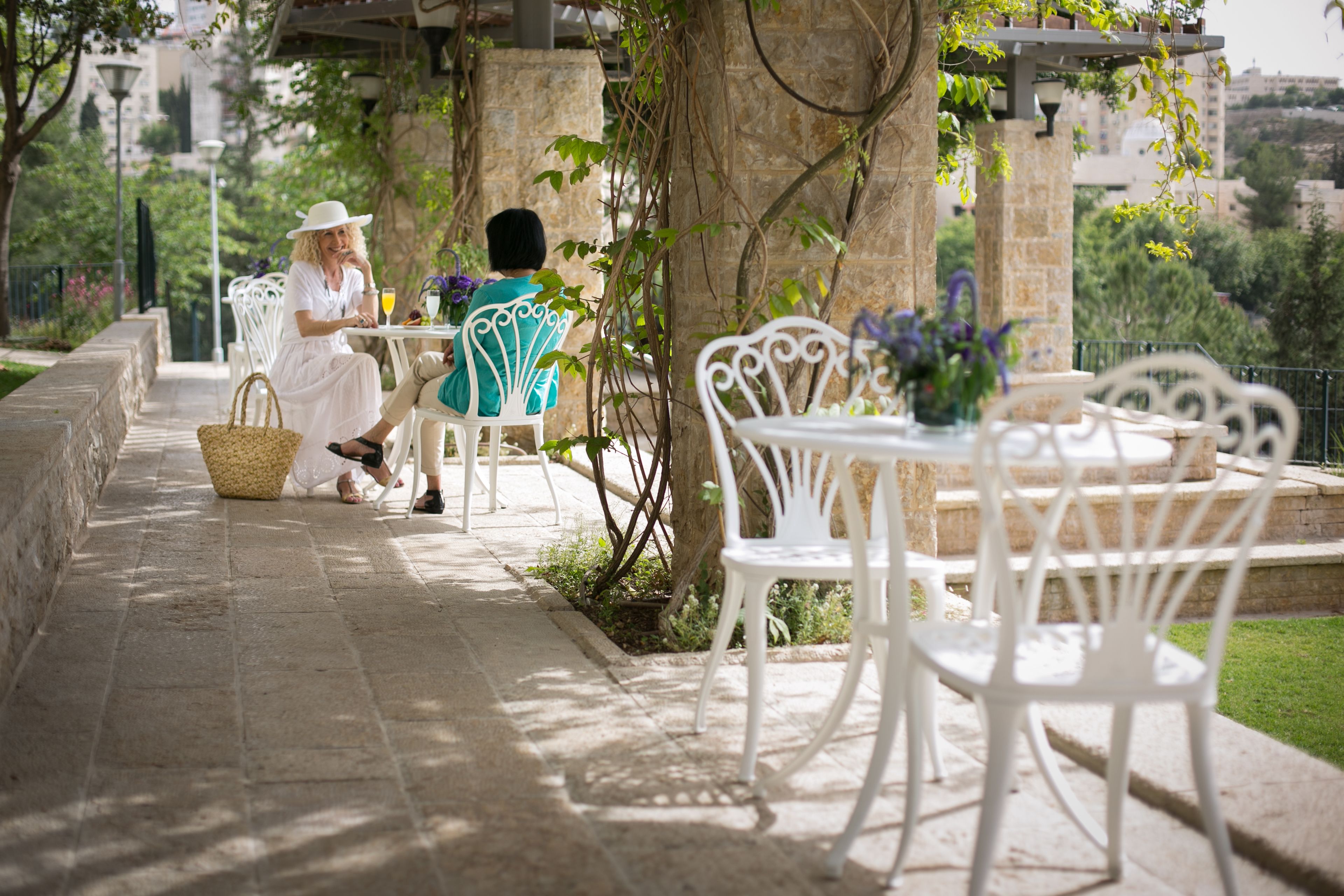 An outdoor seating area with white metal chairs and tables, surrounded by greenery and stone walls.