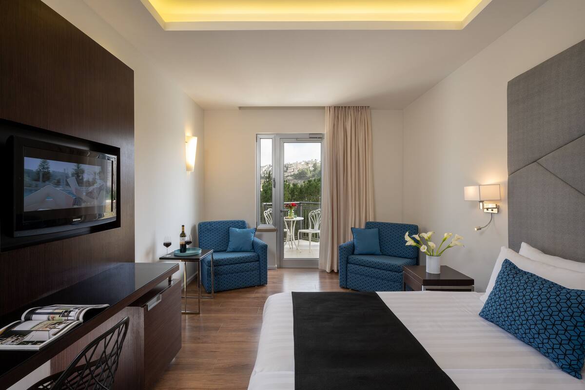 Habitación de hotel con una cama, dos sillones, un televisor, una mesita con una botella y un vaso, y vistas al exterior a través de un ventanal.