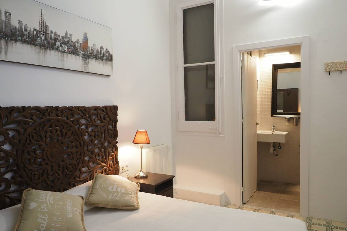 Un dormitorio con cama, cabecera decorativa, lámpara de noche, almohada y un cuadro de paisaje urbano en la pared.