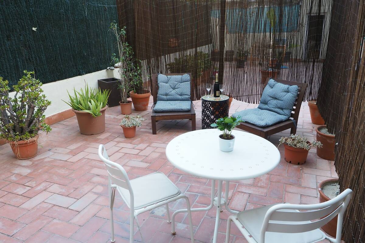 Un patio con una mesa redonda y dos sillas blancas, rodeado de plantas en macetas y una celosía de bambú.