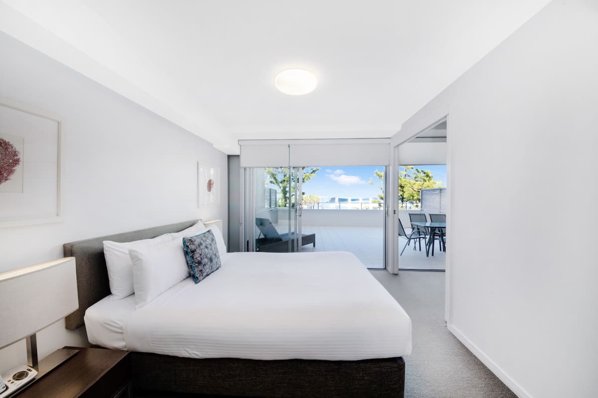 Una habitación de hotel moderna con una cama grande, una mesita de noche y una puerta de cristal que da a un balcón con muebles de exterior.