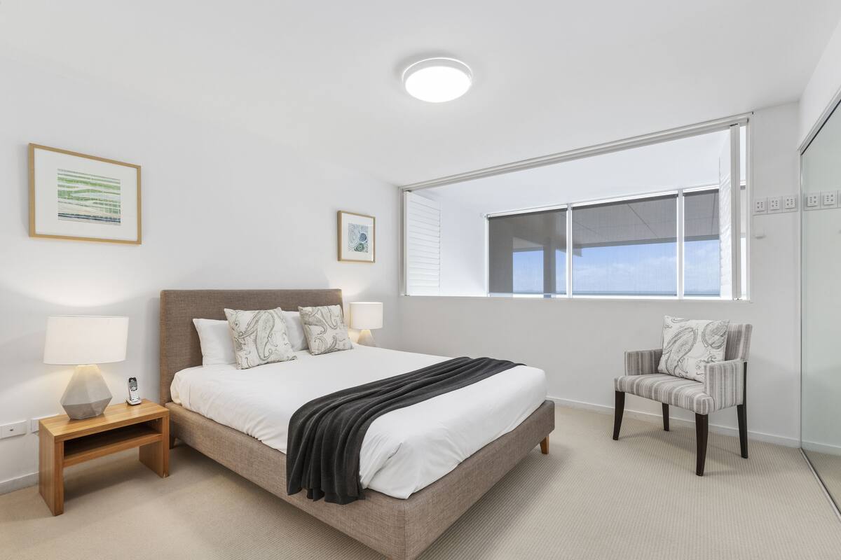 Un dormitorio moderno con una cama grande, mesitas de noche, una silla y una ventana con persianas.