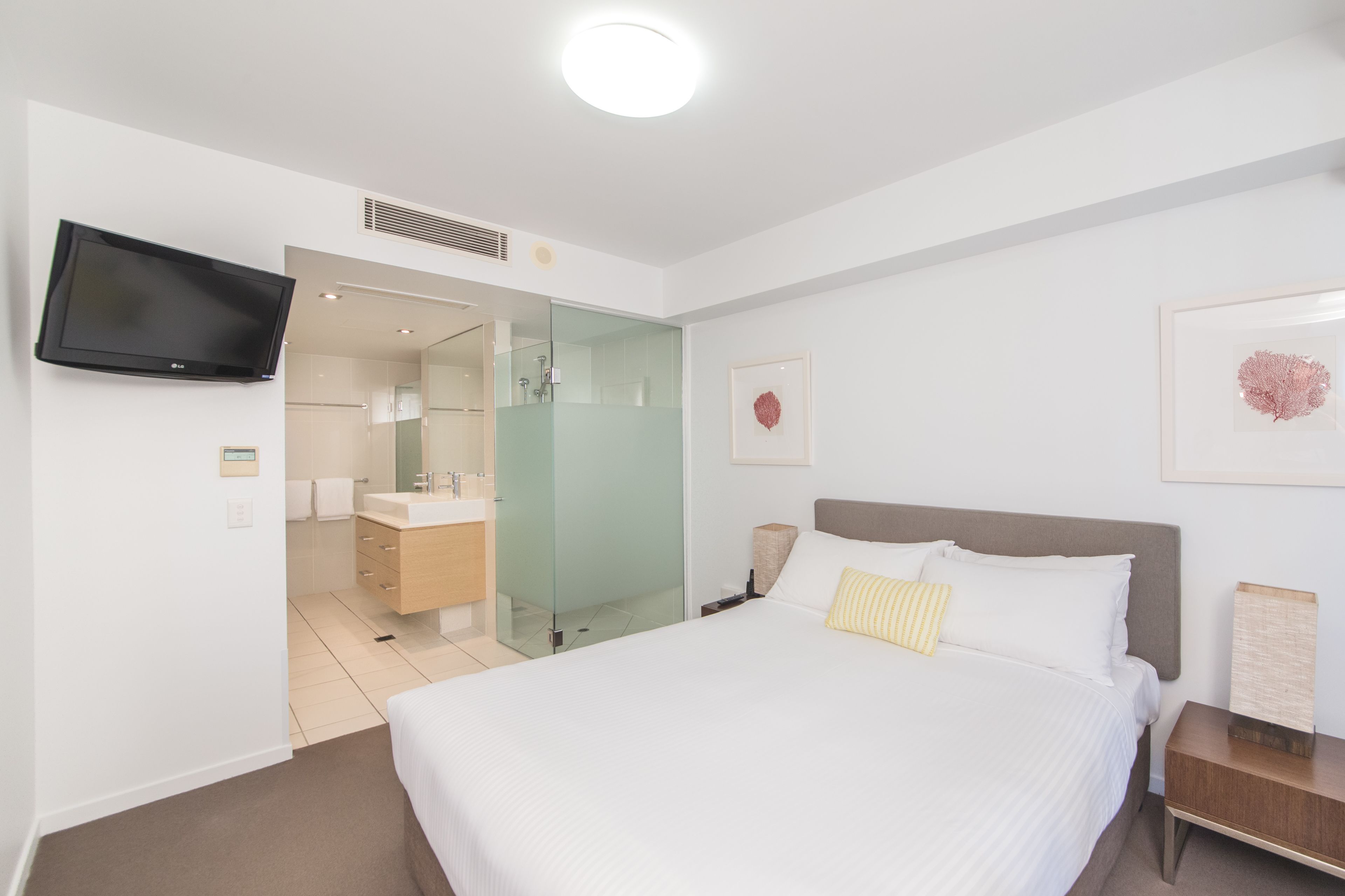 Una habitación de hotel moderna con una cama grande, un televisor montado en la pared, un baño con mampara de vidrio y obras de arte enmarcadas.