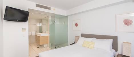 Una habitación de hotel moderna con una cama grande, un televisor montado en la pared, un baño con mampara de vidrio y obras de arte enmarcadas.