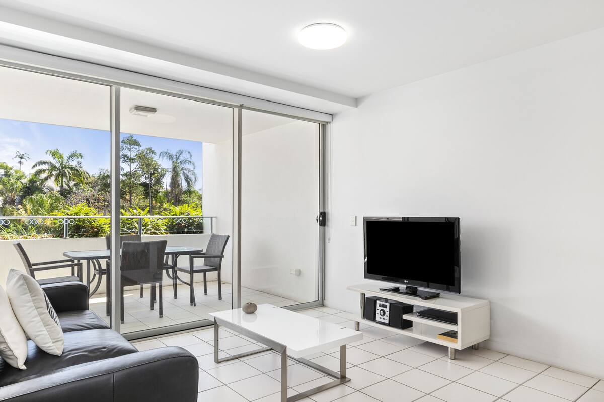 Una sala moderna con sofá, mesa de centro, televisor y puerta corrediza de vidrio que da a un patio exterior con mesa y sillas.