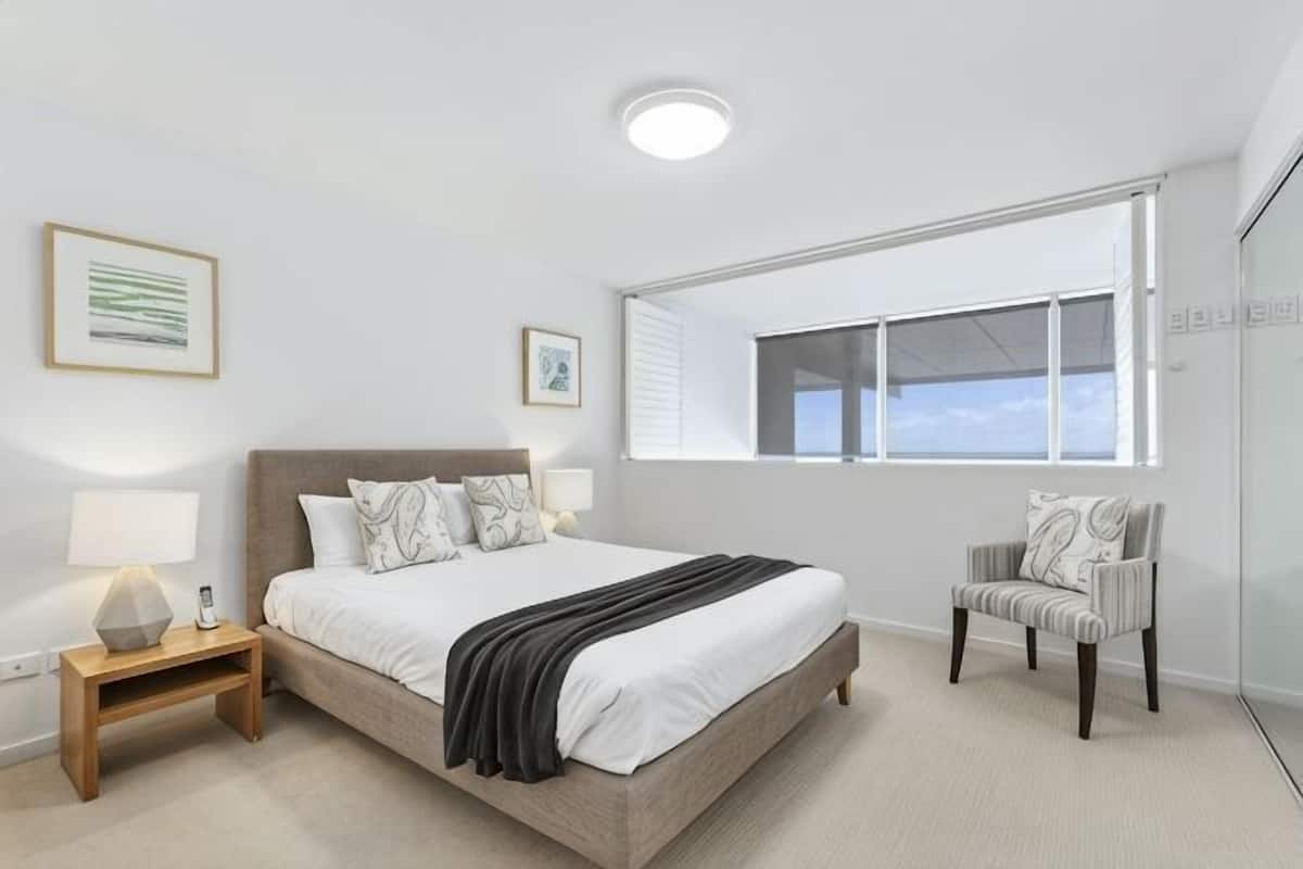 A bedroom with a bed, bedside tables, a chair, a window, and wall art.