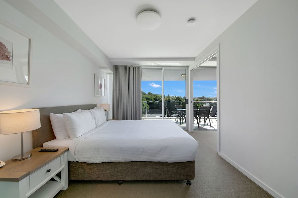 Habitación de hotel con cama, mesita de noche, lámpara y vistas al exterior.