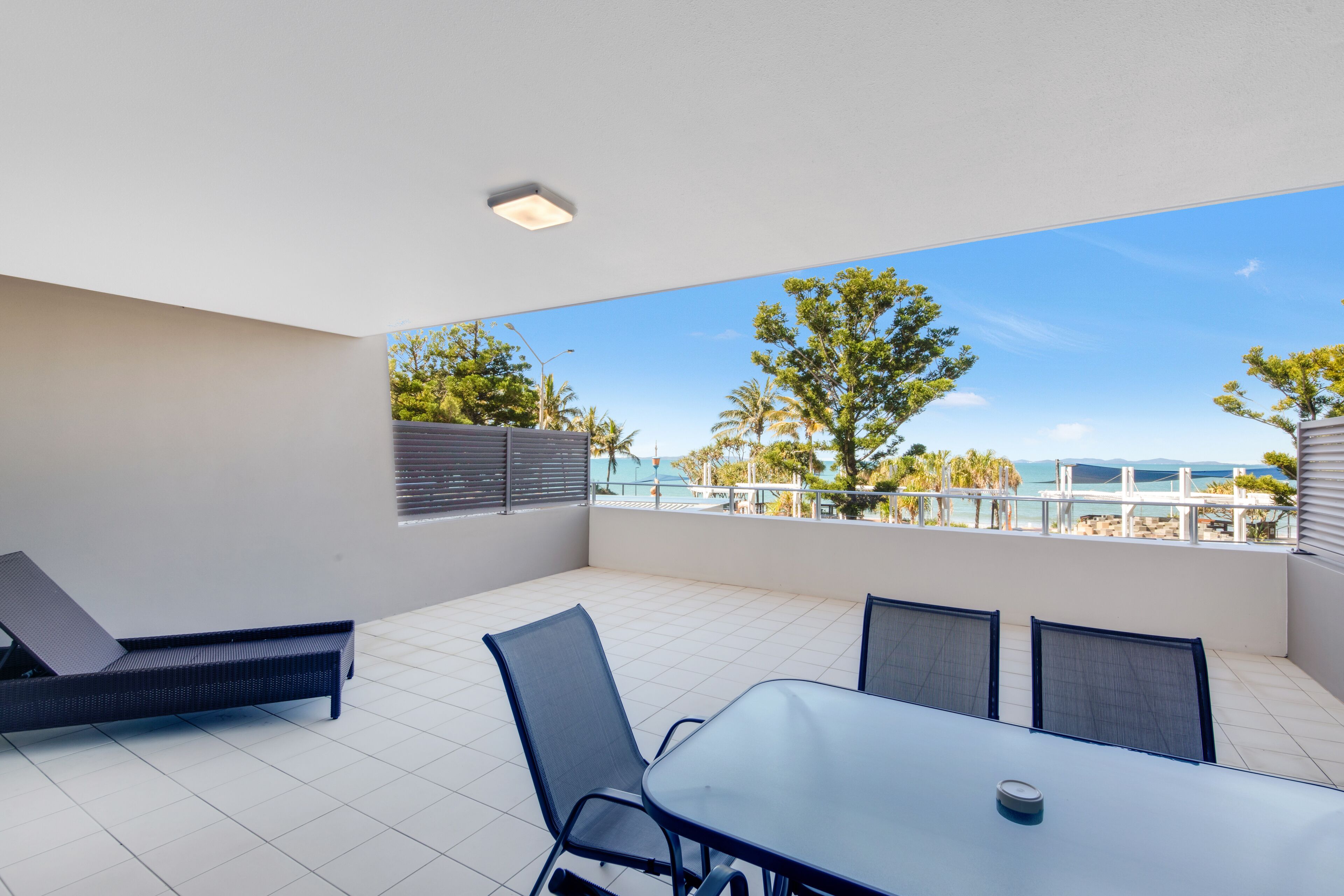 A modern outdoor patio with a dining table and chairs, a lounge chair, and a view of the ocean and palm trees.