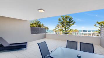 A modern outdoor patio with a dining table and chairs, a lounge chair, and a view of the ocean and palm trees.