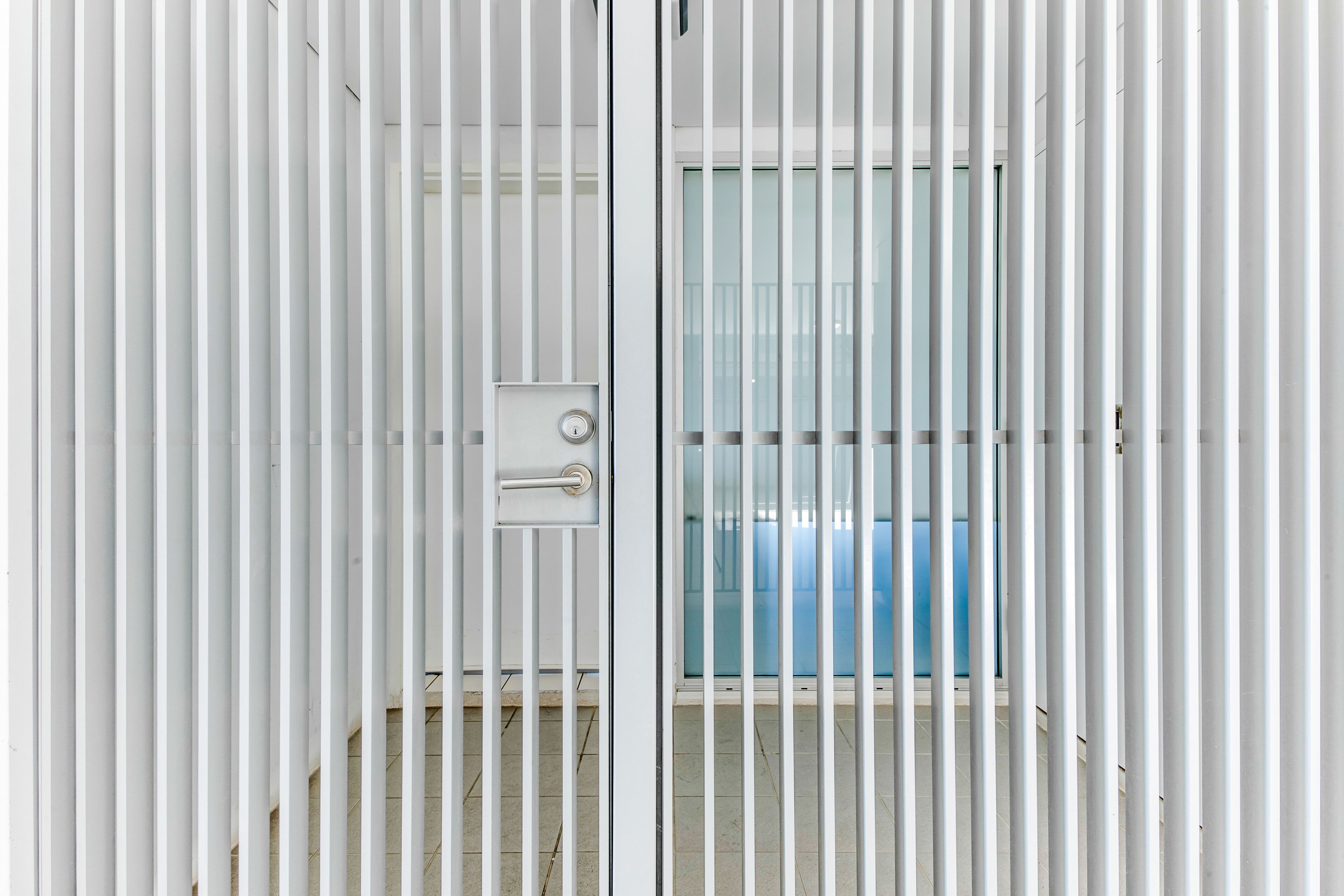 Habitación con persianas verticales blancas, un picaporte visible y una ventana con un objeto azul al fondo.