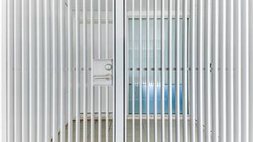 Habitación con persianas verticales blancas, un picaporte visible y una ventana con un objeto azul al fondo.