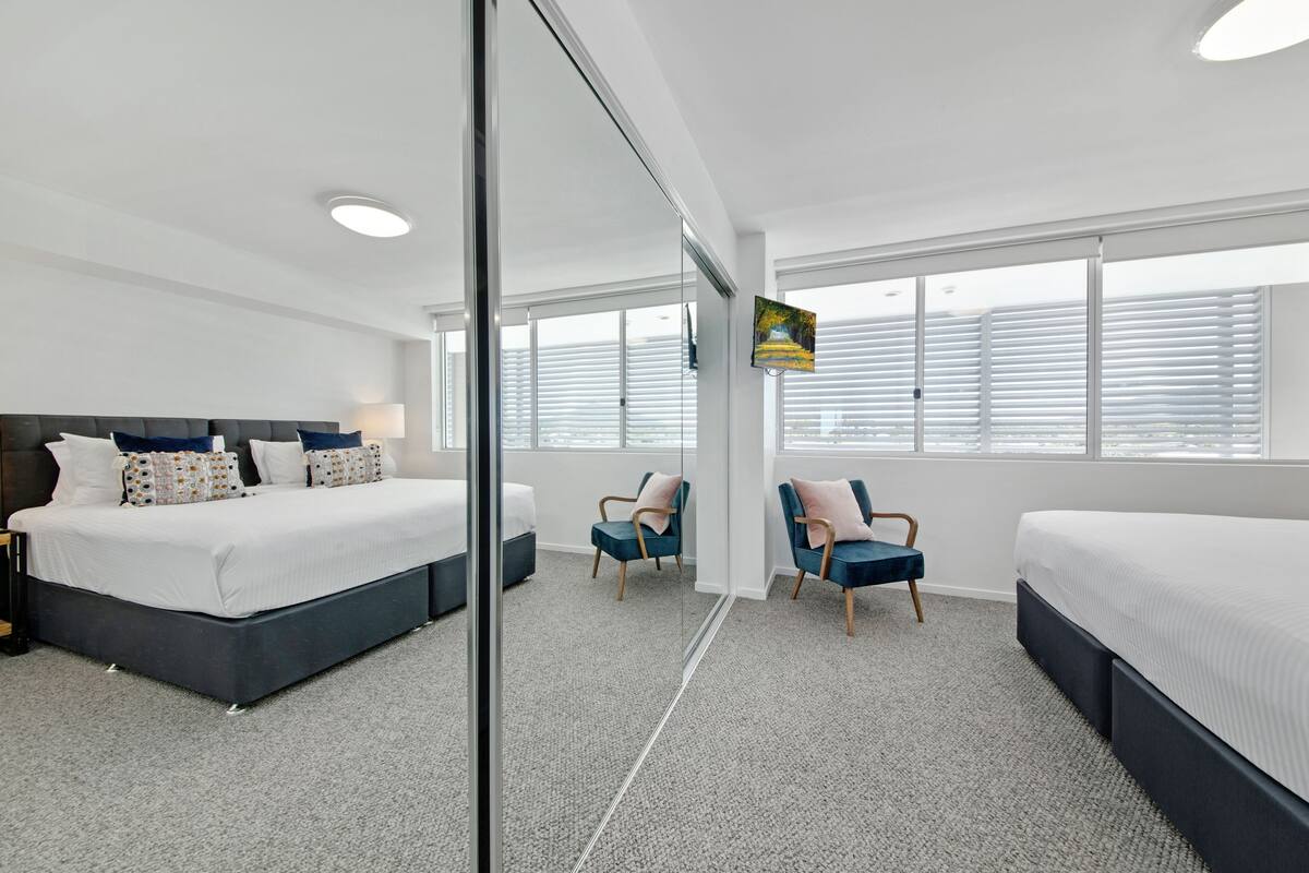 Una habitación de hotel moderna con una cama grande, dos sillas, un televisor y un amplio ventanal con persianas.