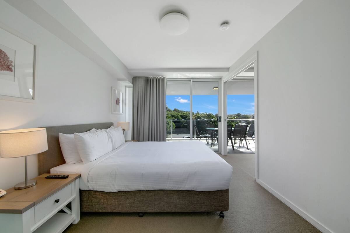 Habitación de hotel moderna con una cama grande, una mesita de noche con lámpara y vistas al patio exterior a través de una puerta corrediza de vidrio.