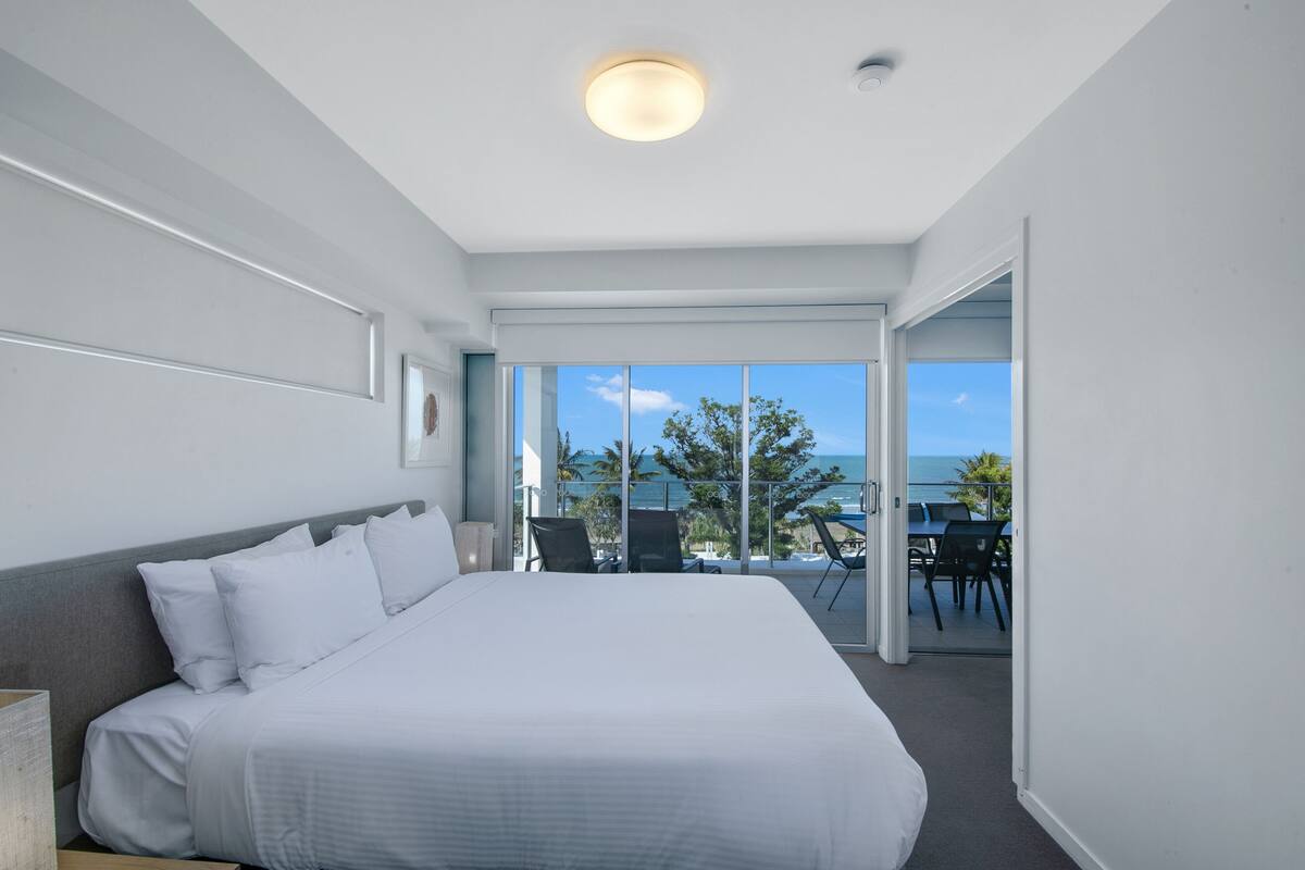 Una habitación de hotel moderna con una cama grande, vista al océano y un balcón con mobiliario exterior.