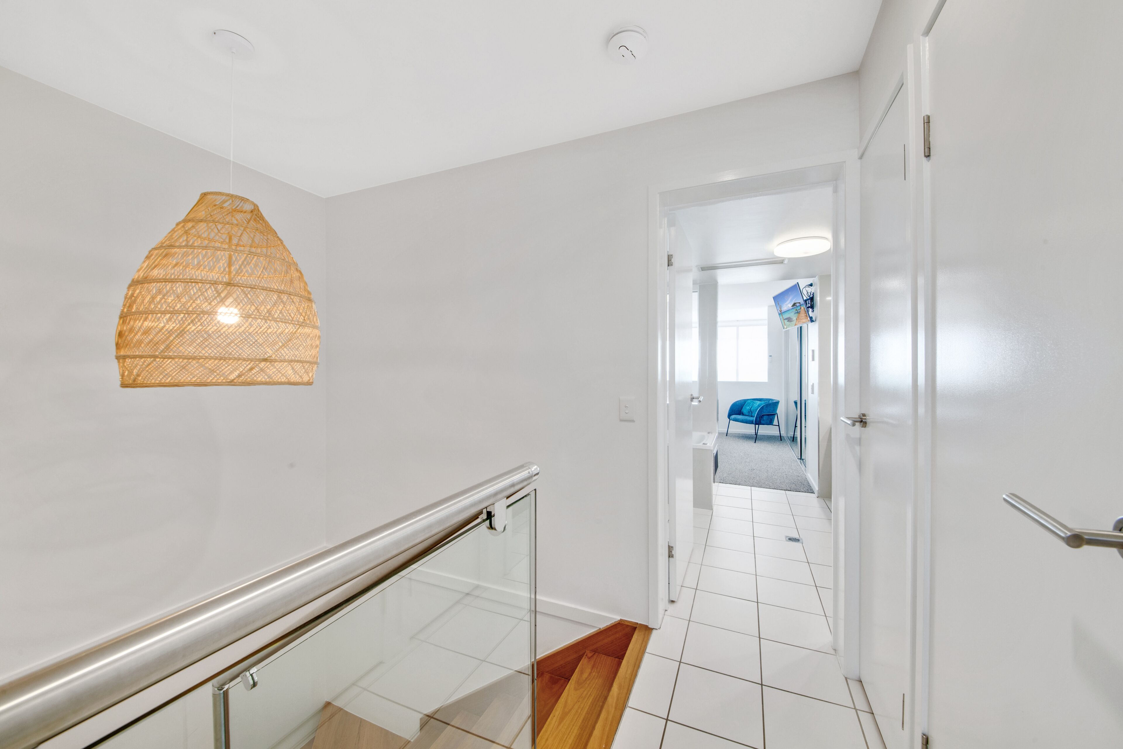 Een modern interieur met glazen balustrade, een geweven lamp en een betegelde vloer die leidt naar een ruimte met een blauwe stoel.