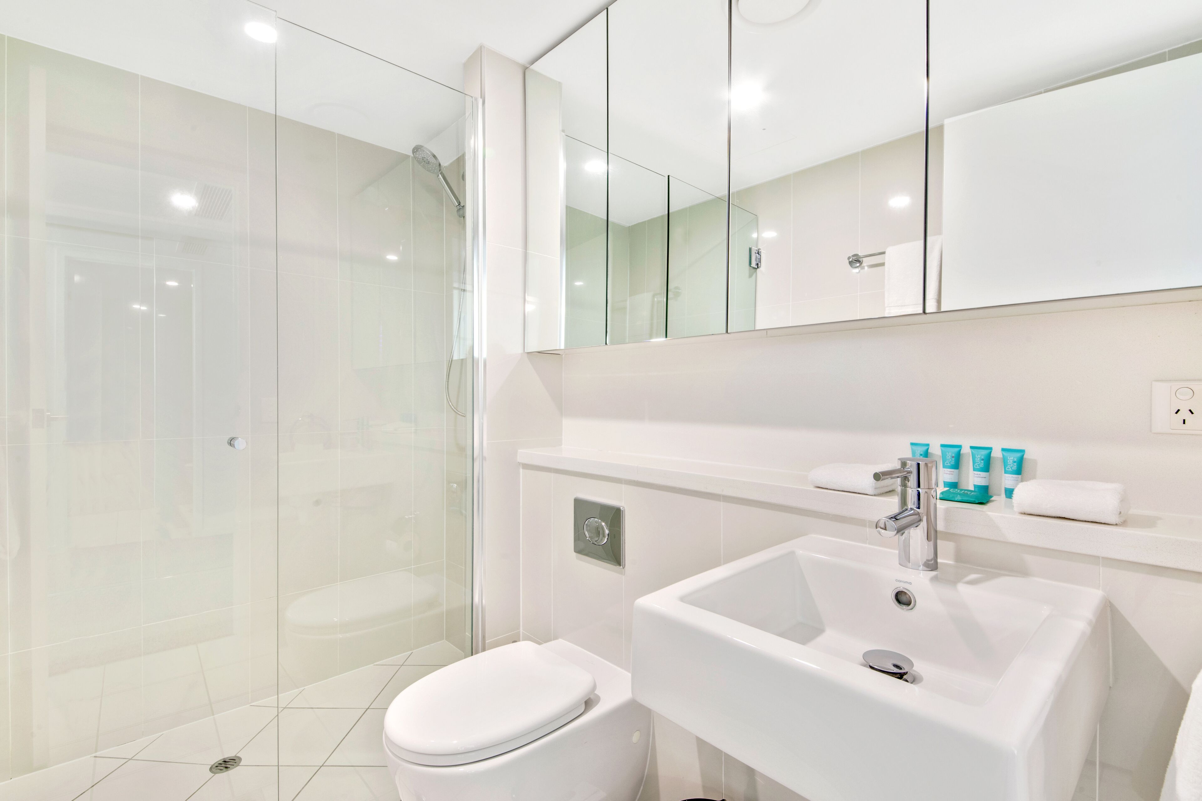 Une salle de bain moderne avec une douche à parois de verre, des toilettes blanches et un lavabo surmonté d’un miroir.
