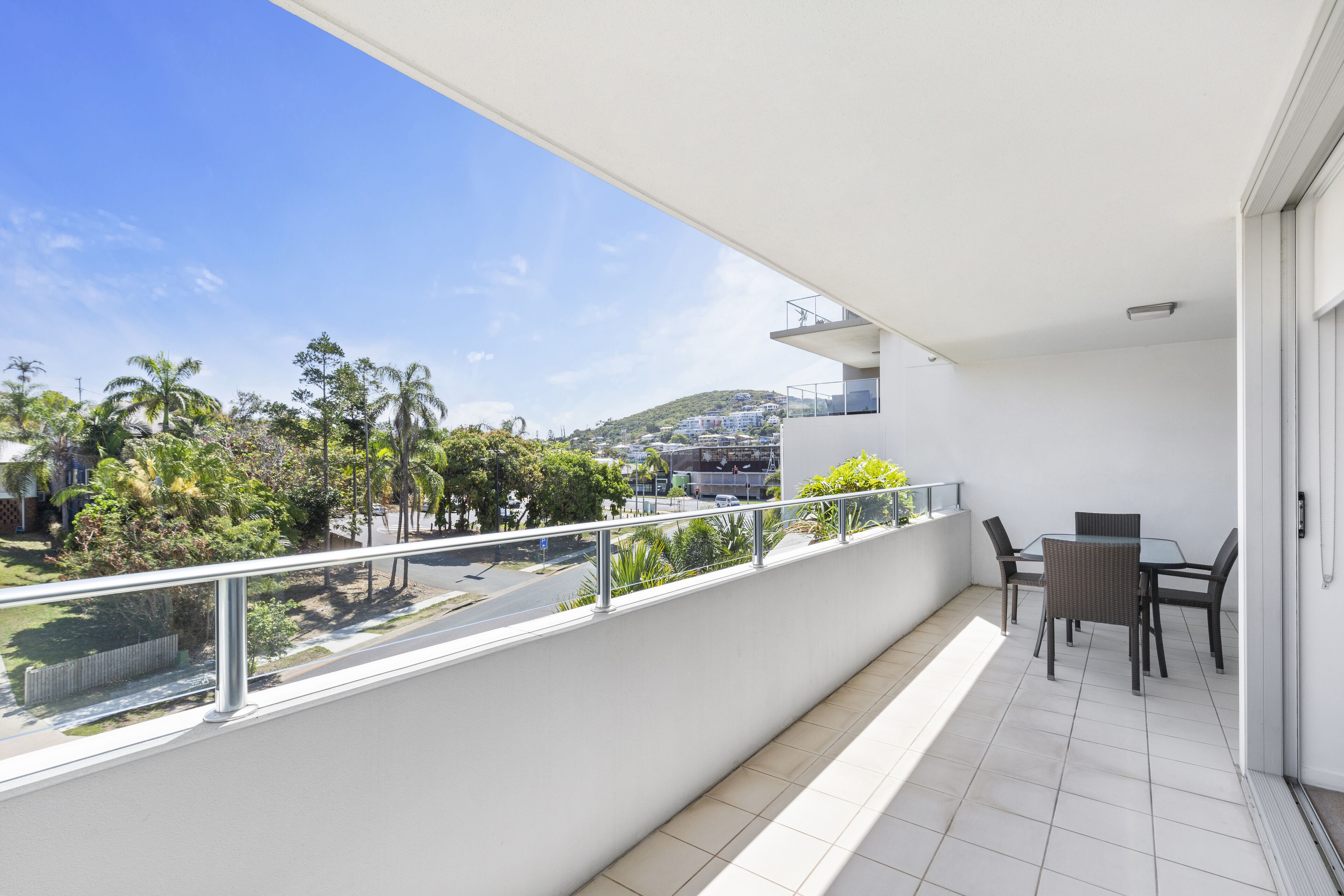 A balcony with a table and chairs, a white railing, and a view of a green landscape with trees and buildings in the distance.