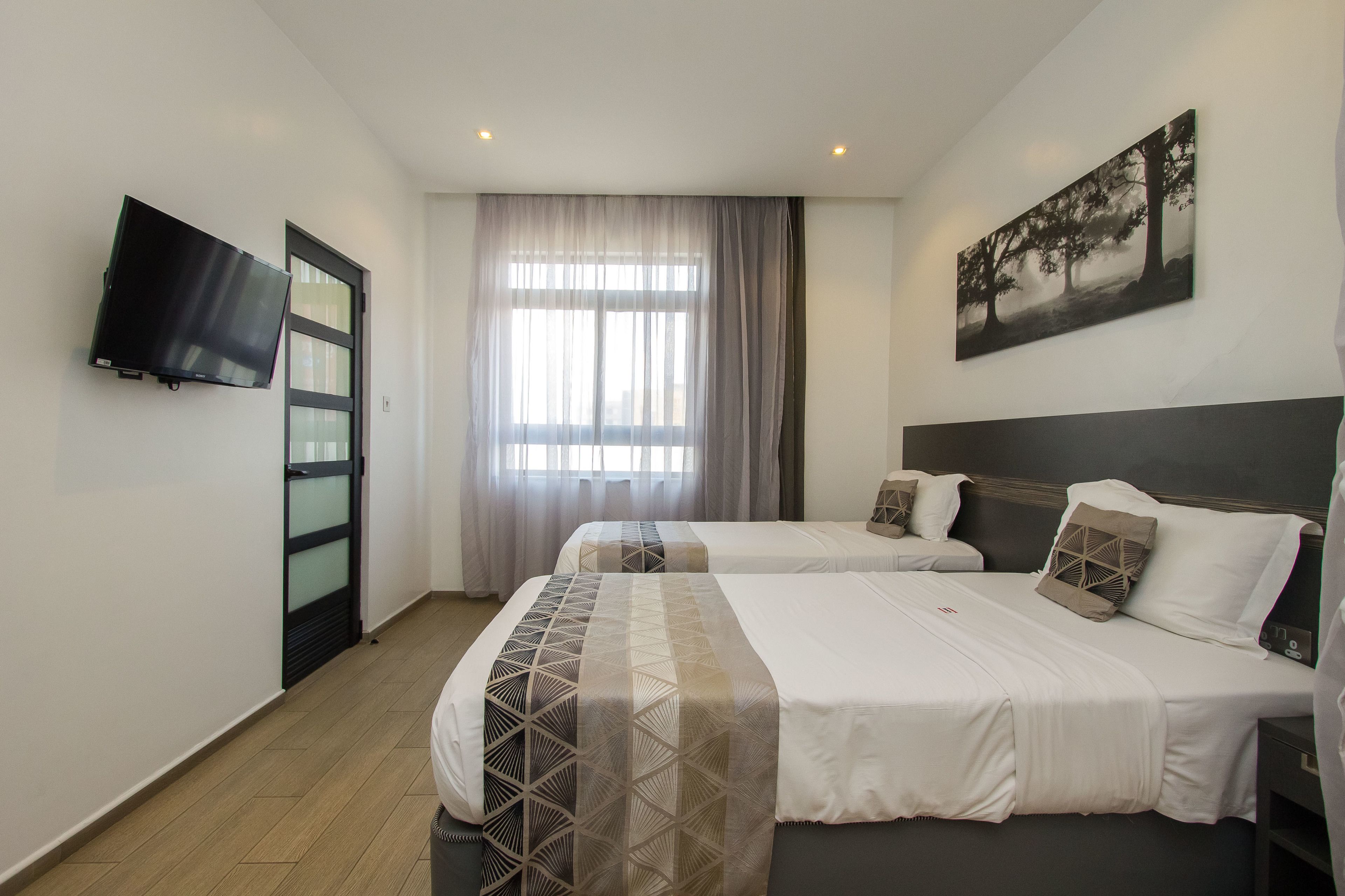 A hotel room with two beds, a flat-screen TV mounted on the wall, a window with sheer curtains, and a painting on the wall.