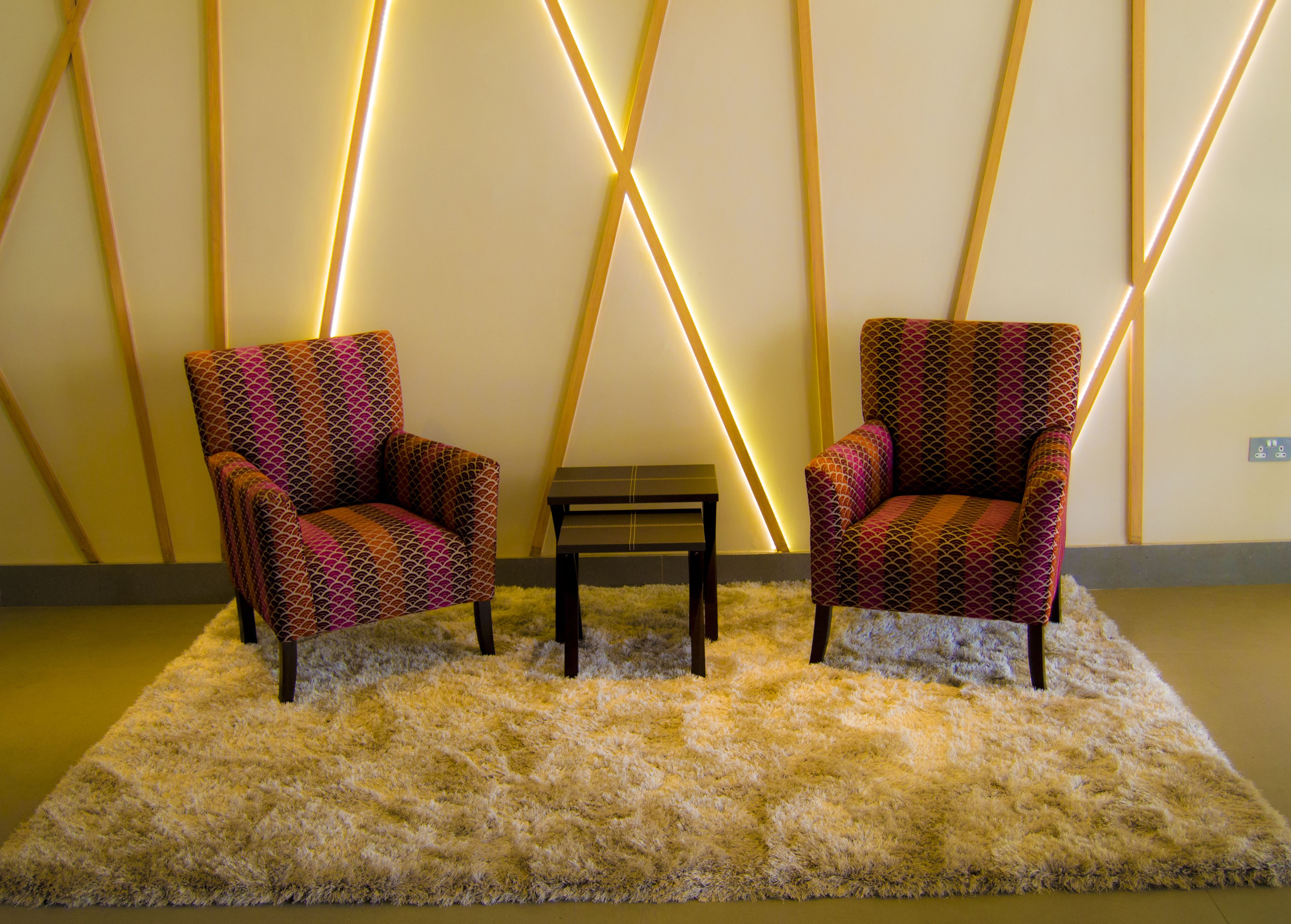 Two patterned armchairs with a small table in between, set on a fluffy rug against a wall with geometric lighting.