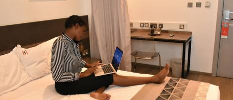 A person sitting on a bed with a laptop, a remote control, and a desk with a telephone and a cup in the background.