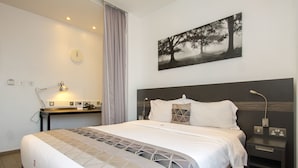 A hotel room with a large bed, a desk with a lamp, a clock, and a painting of trees. - Imperial Hotel Express (Kisumu)