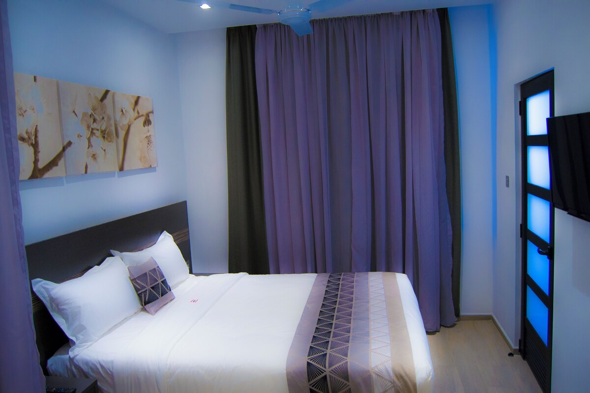 A hotel room with a bed, curtains, a ceiling fan, and artwork on the wall.