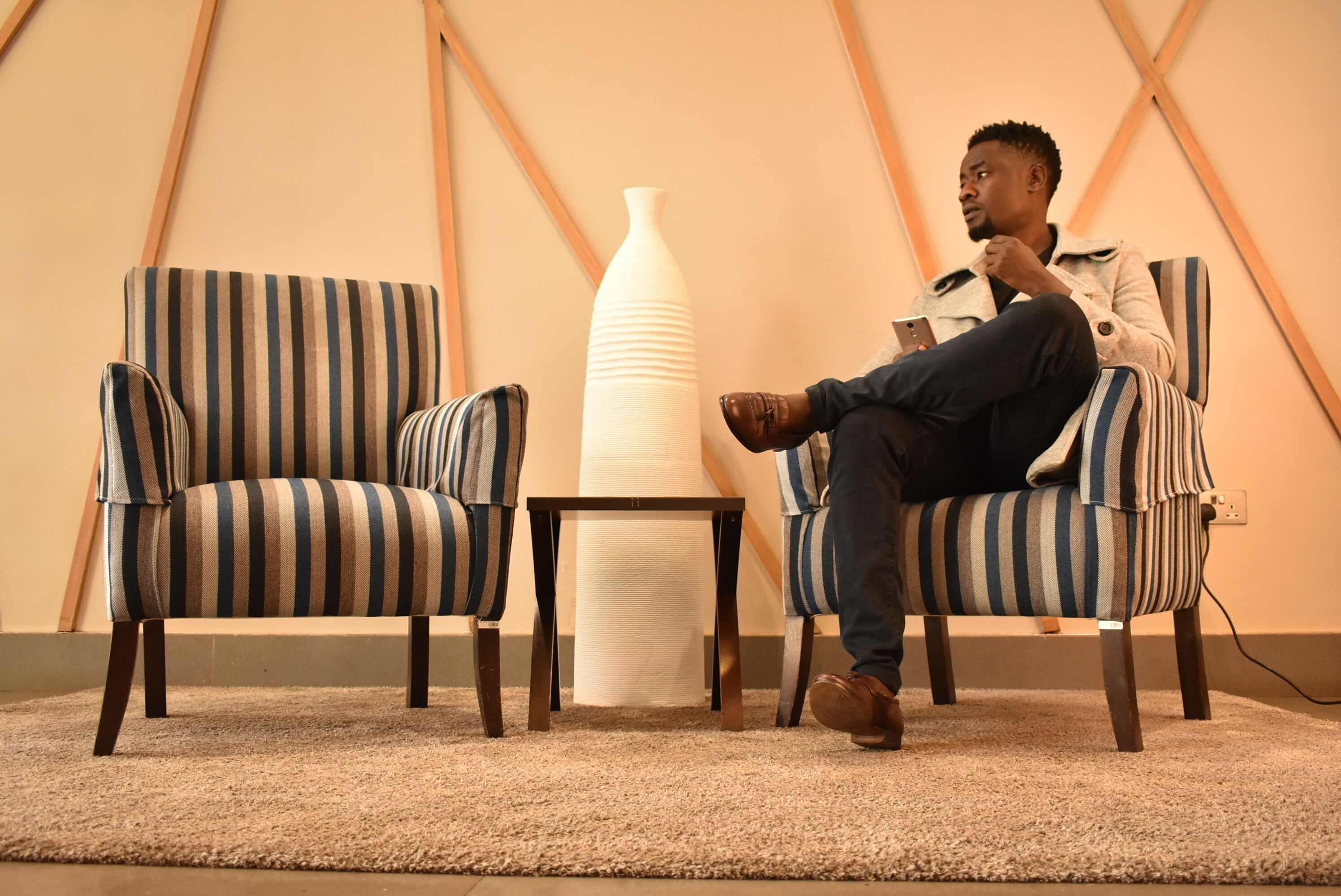 A person sitting on a striped chair in a room with a large vase and a small table.