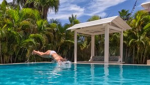 Une piscine où une personne est en train de plonger, entourée de palmiers et d’un espace de repos abrité.