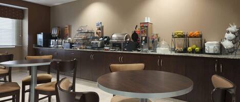 A hotel breakfast area with round tables and chairs, a coffee station, and a fruit display.