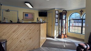 A reception area with a wooden reception desk, framed certificates, an American flag, a large mirror, and a view through a window. - KC Motel (Show Low)
