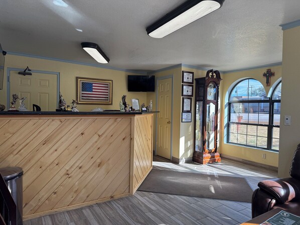 A reception area with a wooden reception desk, framed certificates, an American flag, a large mirror, and a view through a window. - KC Motel (Show Low)