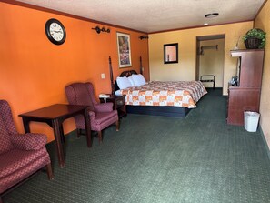 A hotel room with a bed, a desk, a chair, a nightstand, a clock, and a trash bin. - KC Motel (Show Low)