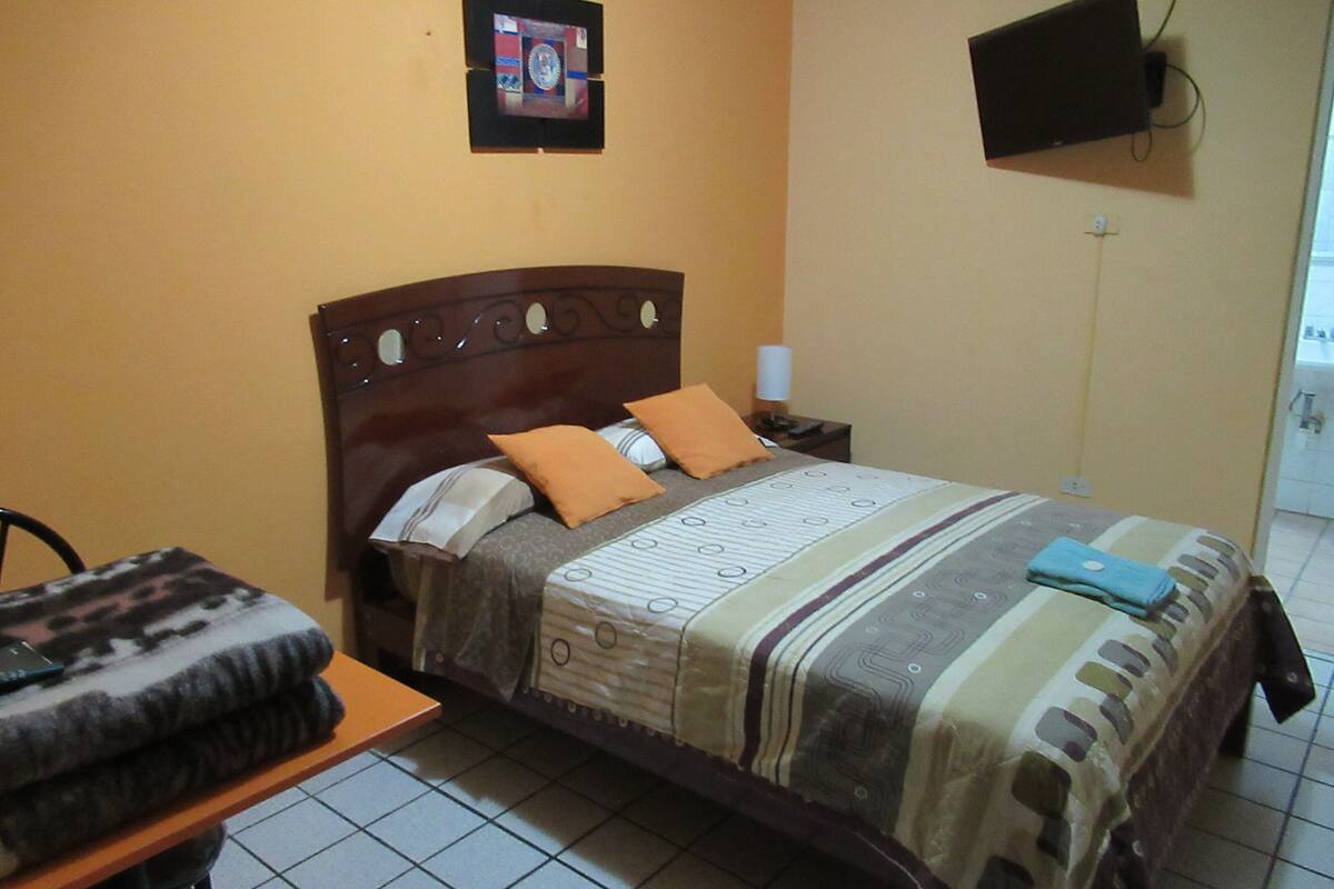 Une chambre d’hôtel avec un lit, une table de chevet, un téléviseur fixé au mur et une petite table avec une couverture.