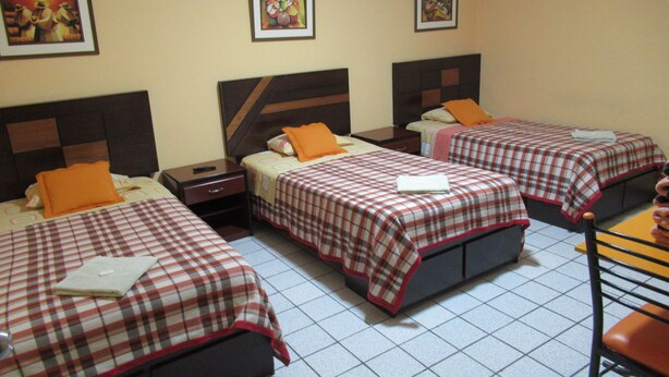 Habitación de hotel con tres camas, cada una con una colcha a cuadros y un almohá de naranja. Hay cuadros enmarcados en la pared sobre las camas.