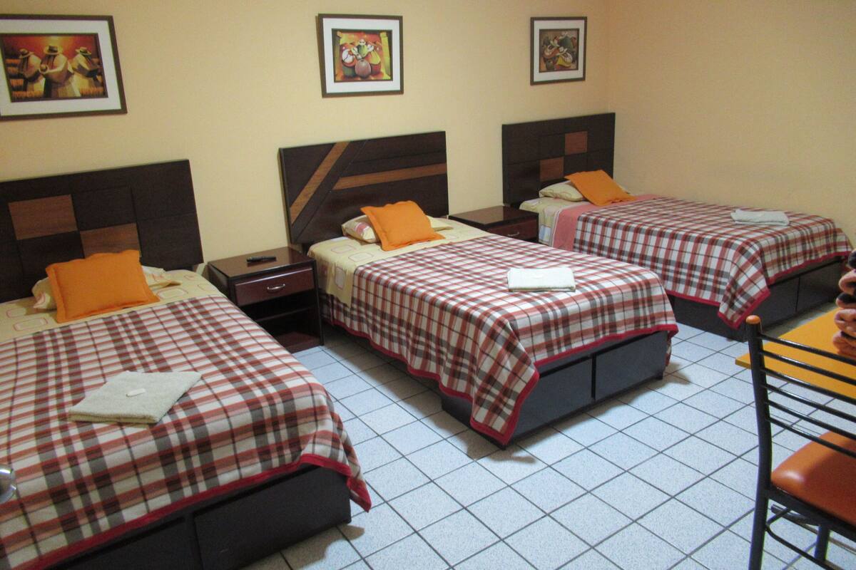 Une chambre d’hôtel avec trois lits, chacun recouvert d’une courtepointe à carreaux et agrémenté d’un oreiller orange. Des tableaux encadrés sont accrochés au mur au-dessus des lits.