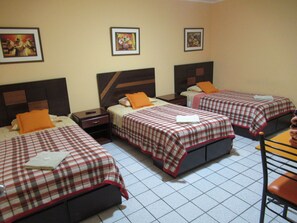 A hotel room with three beds, each with a checkered bedspread and an orange pillow. There are framed pictures on the wall above the beds. - Kurmi Hostel Lima Airport (Callao)