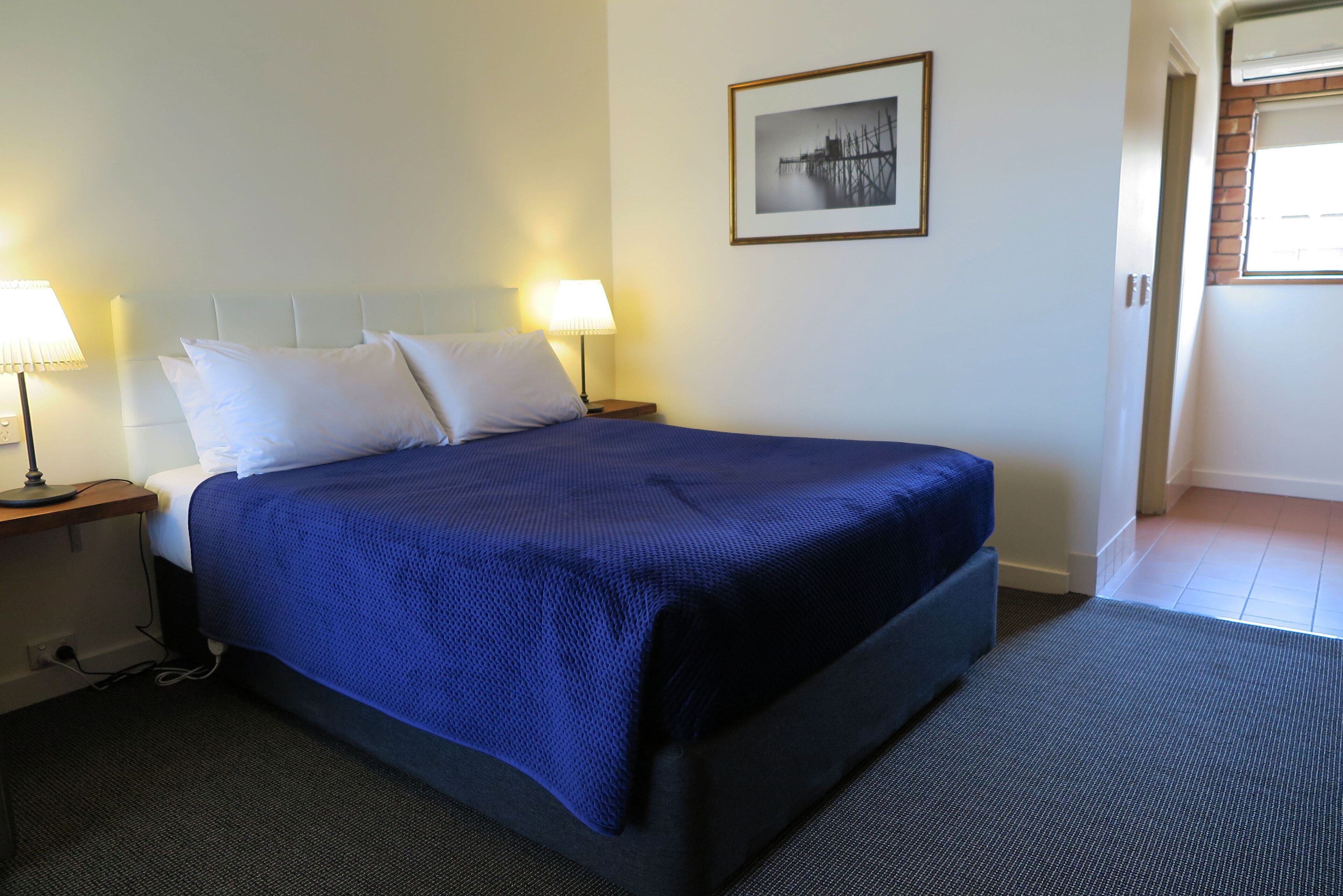 A hotel room with a large bed, two bedside lamps, a framed picture on the wall, and a view of the outside through a window.