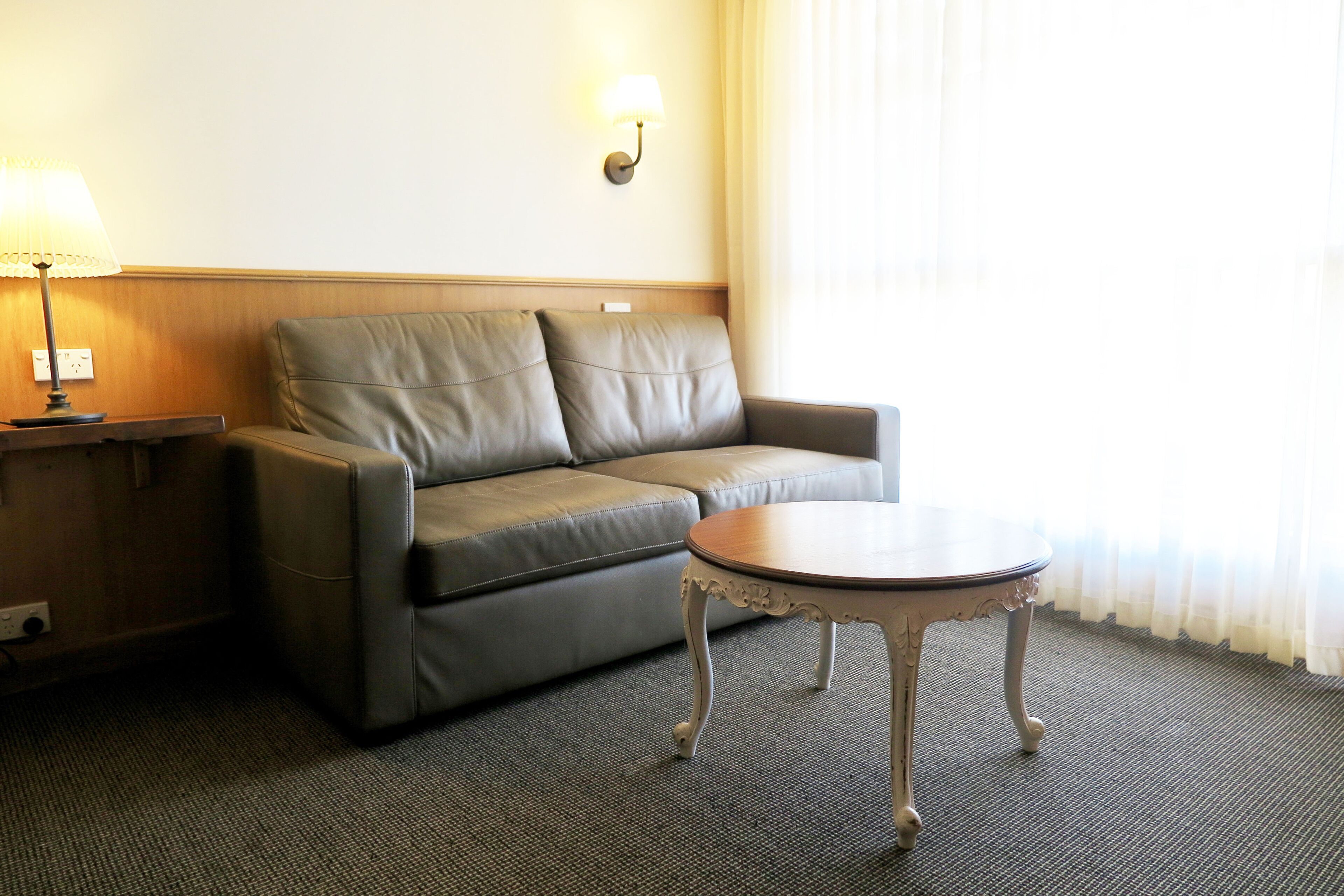 A room with a grey leather sofa, a small round table, a lamp, and a wall-mounted light fixture.