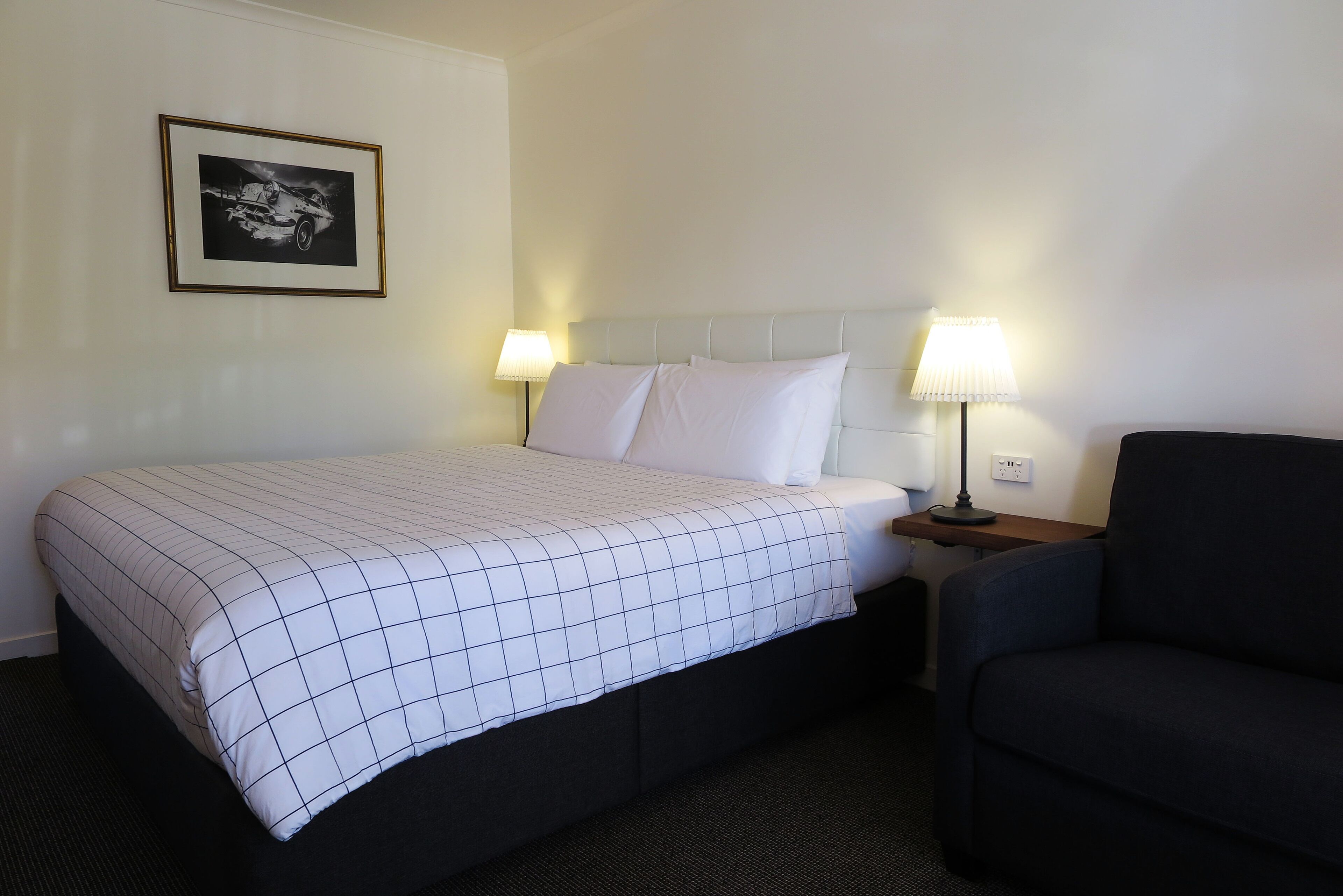 Una habitación de hotel con cama, dos lámparas de noche, un cuadro enmarcado en la pared y un sillón de color oscuro.