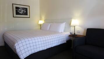 Una habitación de hotel con cama, dos lámparas de noche, un cuadro enmarcado en la pared y un sillón de color oscuro.