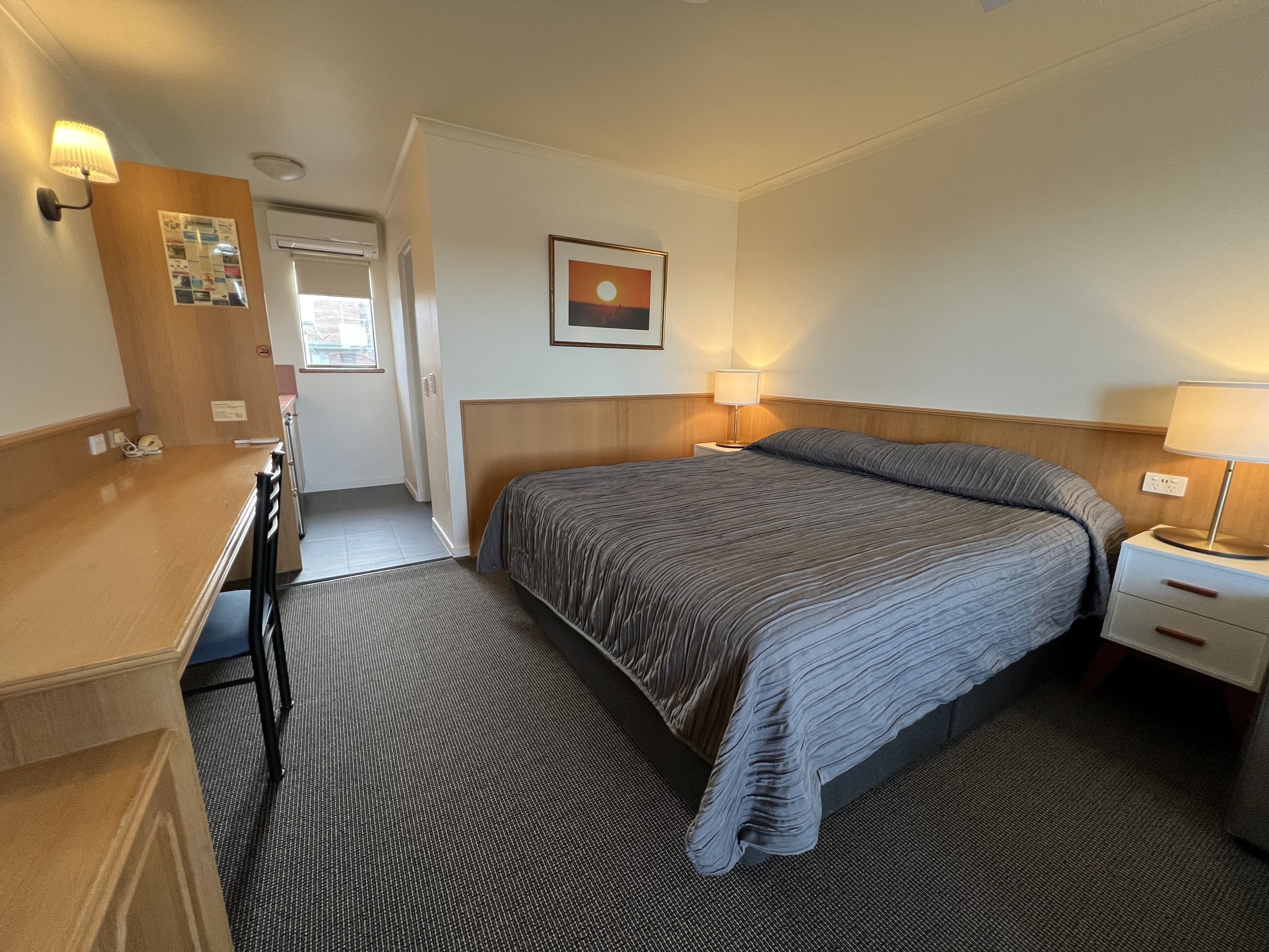 Et hotelværelse med seng, sengeborde, et skrivebord og en lampe på væggen.