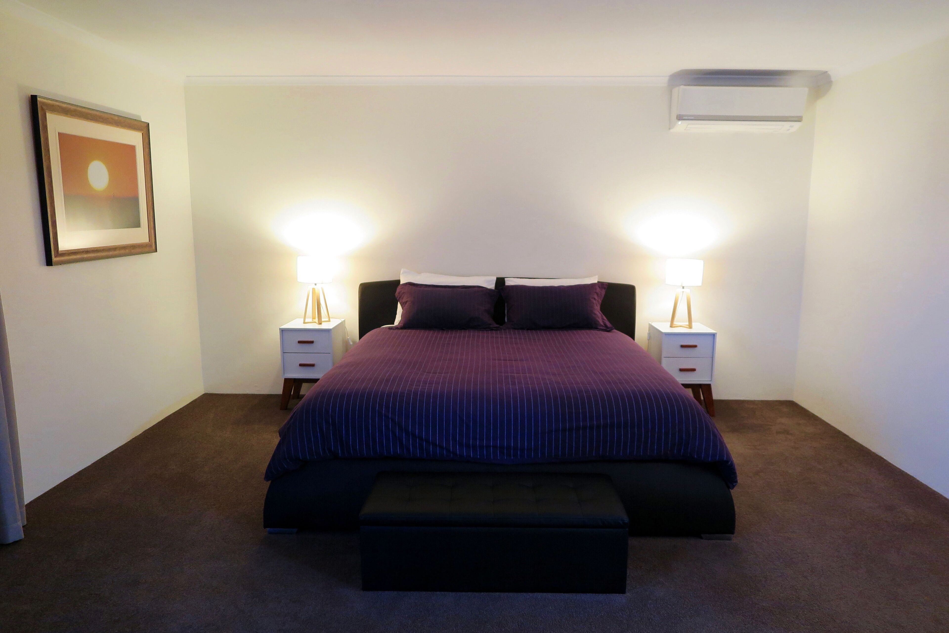 Un dormitorio con una cama grande, dos mesitas de noche, un buró, un aire acondicionado de pared y un cuadro enmarcado en la pared.